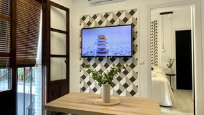 Ein modernes Zimmer mit einem an der Wand mit geometrischem Muster befestigten Fernseher, einem Holztisch mit einer Vase und Blick durch ein mit Jalousien versehenes Fenster nach draußen.
