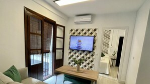 Ein modernes Wohnzimmer mit einer Couch, einem Couchtisch aus Holz mit einer Vase, einem an einer Wand mit geometrischem Muster montierten Fernseher und einer Schiebetür zu einem Badezimmer.
