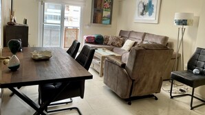 Ein Wohnzimmer mit einem braunen Sofa, einem Esstisch und einem Couchtisch. Ein Fenster mit Jalousien, ein Bild an der Wand und eine Stehlampe sind ebenfalls vorhanden.