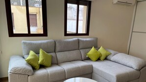 A grey sectional sofa with green cushions, a white round coffee table, and two windows with blinds. - Casa Terraza Judería: Spacious History-Filled Retreat with Private Terrace in Córdoba's Jewish Quarter (Córdoba)