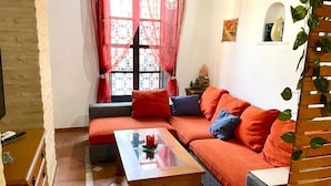 Ein gemütliches Wohnzimmer mit einem Ecksofa, einem Holztisch und einem Fenster mit gemusterten Vorhängen.