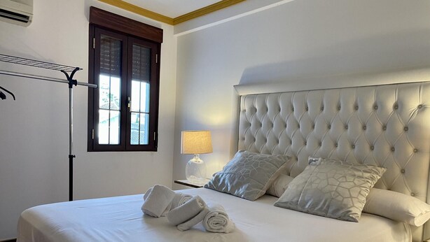 Uma cama bem arrumada com lençóis brancos e toalhas dobradas, uma cabeceira estofada e um abajur.