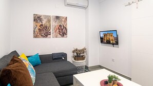 Un salon moderne avec un canapé gris, des coussins colorés, une table basse blanche agrémentée de plantes, un téléviseur fixé au mur et un climatiseur mural.