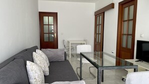 Ein modernes Wohnzimmer mit grauem Sofa, einem Glastisch, einem Fernseher und einem kleinen weißen Tisch mit Stühlen.