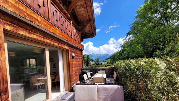 A wooden building with a glass door, outdoor seating, and a view of mountains.