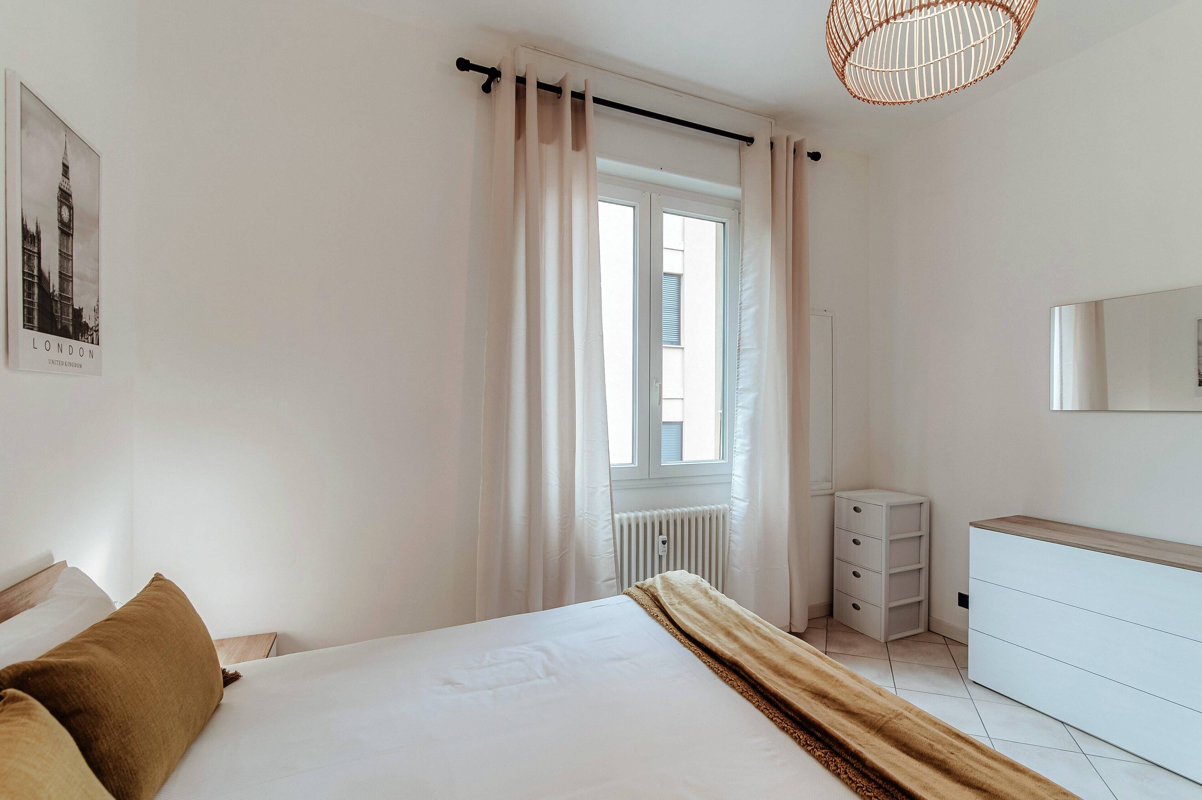 Une chambre à coucher avec un lit, une commode, un radiateur et une fenêtre avec des rideaux.