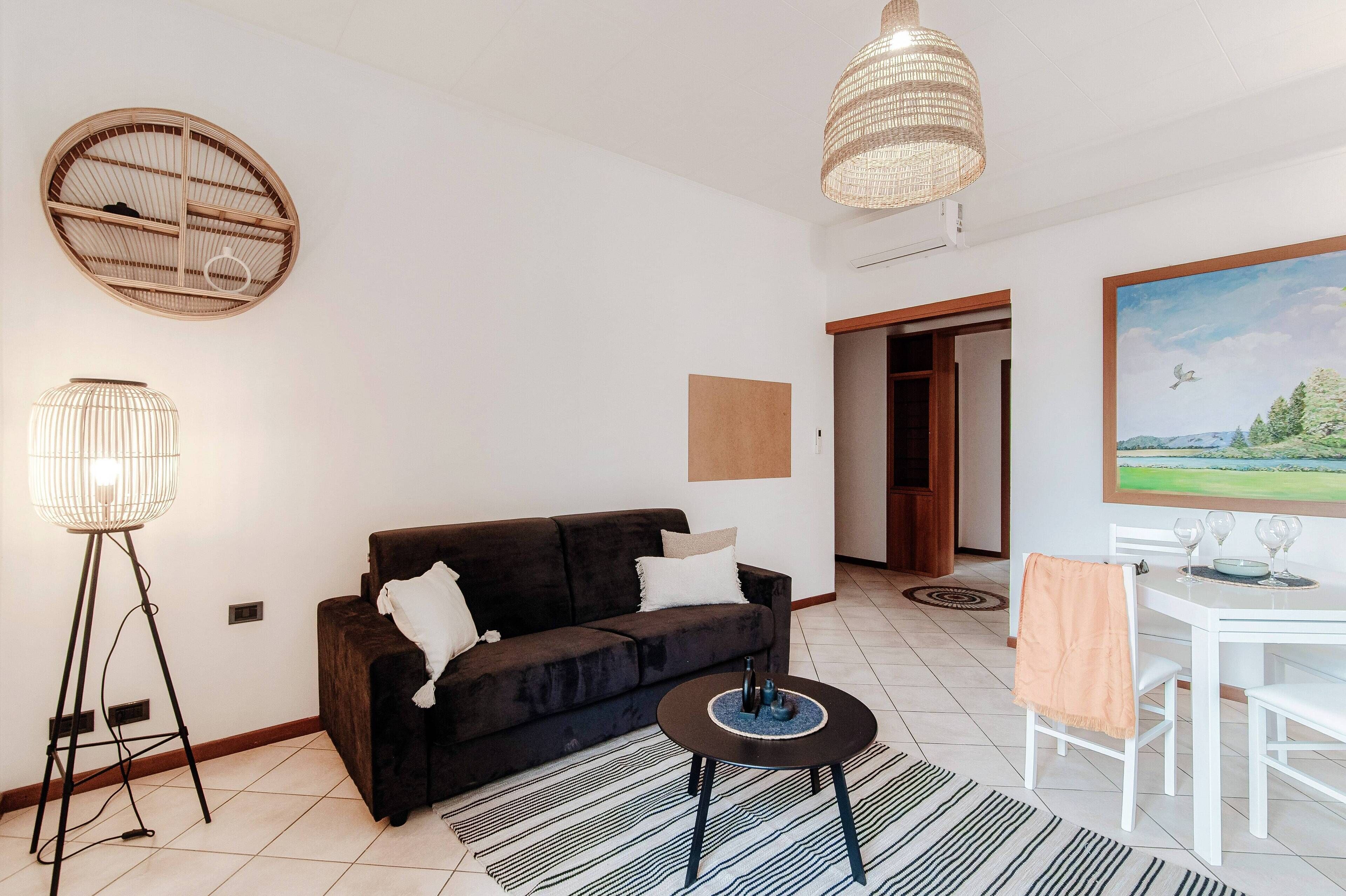Un salon moderne avec un canapé foncé, une table basse ronde, un lampadaire, un luminaire suspendu et un coin repas avec une table et des chaises blanches.