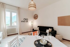 A modern living room with a white table set for dining, a black sofa, and a large window with curtains. - Valli (Como)