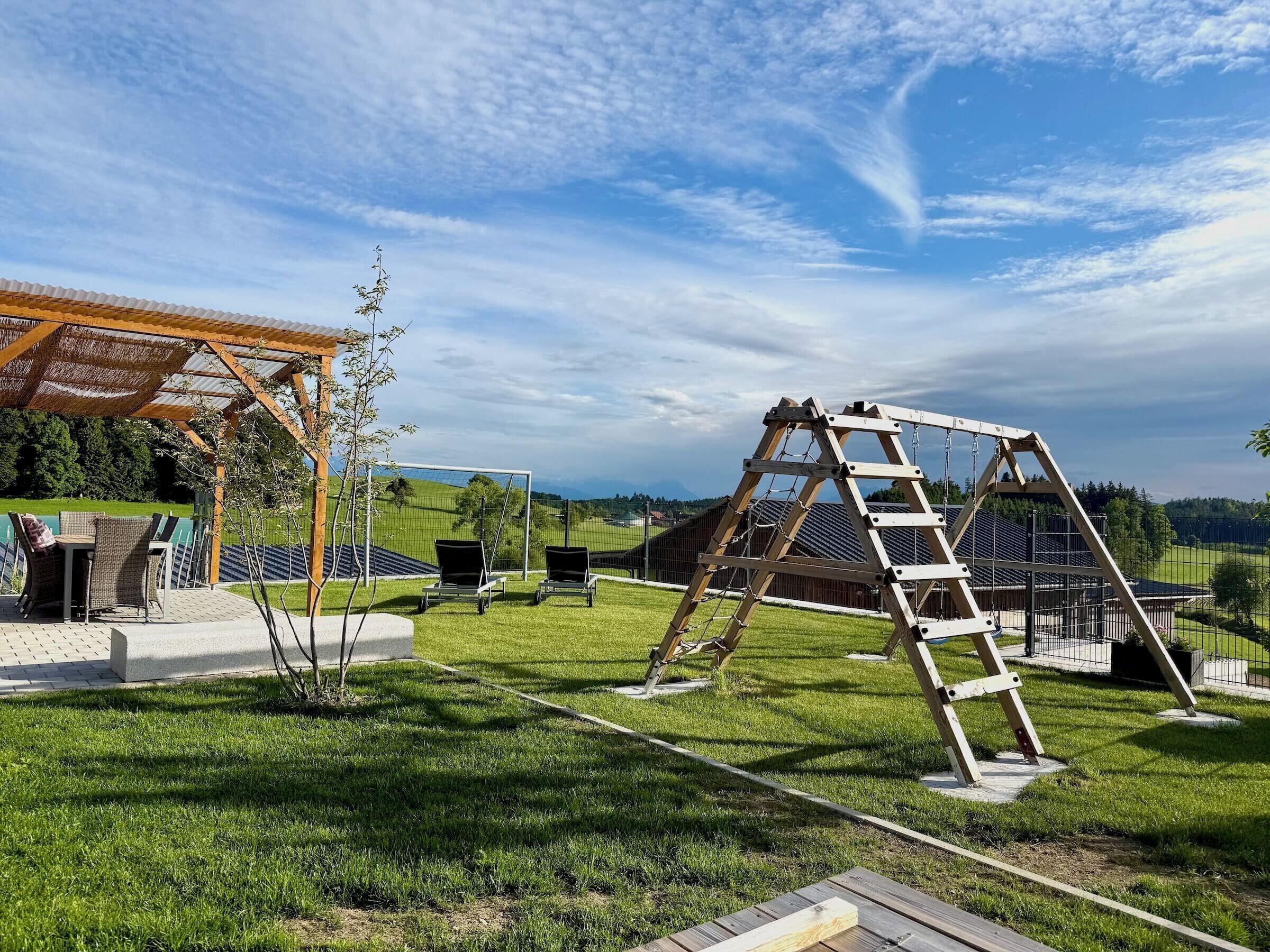 A grassy outdoor area with a wooden swing set, a covered seating area, and a view of distant hills.