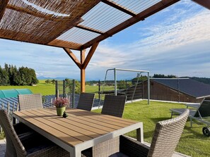 Eine Außenpatio mit einem Holztisch und Korbstühlen, einer Pergola mit Mesh-Dach und Blick auf ein Sportfeld und Berge im Hintergrund.