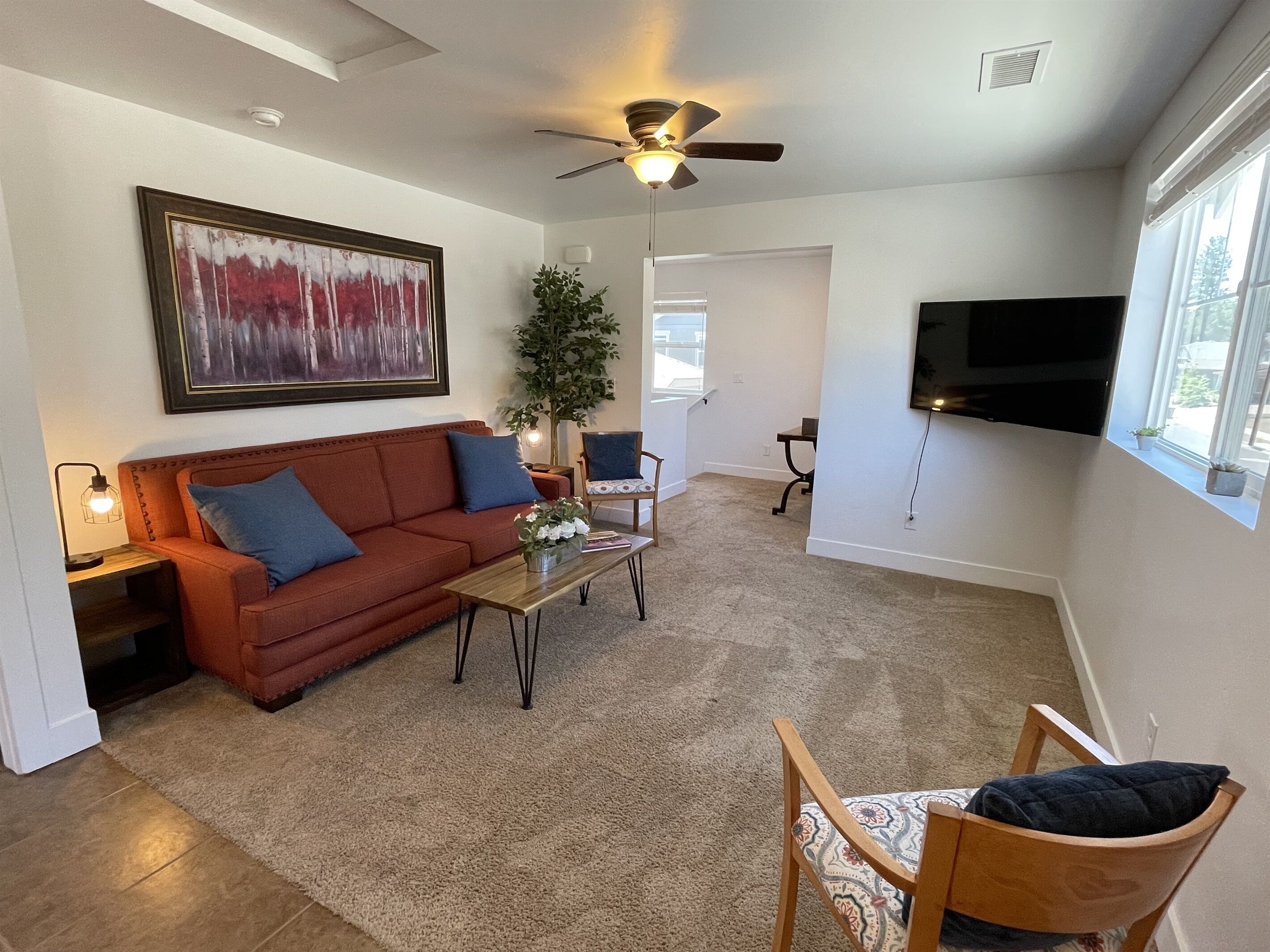 Un salon avec un canapé, une table basse et des fauteuils. On y trouve un ventilateur de plafond, un téléviseur fixé au mur et une plante.