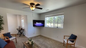 A living room with a ceiling fan, a flat-screen TV, two chairs, a coffee table, and a window with blinds. - Windtree Hideaway! Trees, hiking, minutes downtown (Flagstaff)