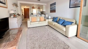 Ett modernt vardagsrum med en beige soffa, en platt-TV och ett kök i bakgrunden.