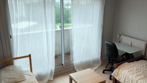 A hotel room with two beds, a small table, a chair, and a view of a tennis court through sheer curtains.