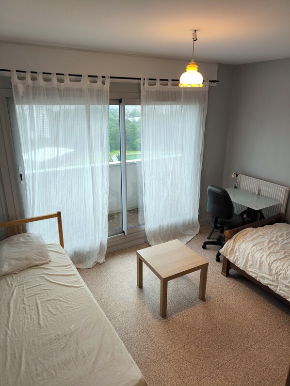 Een hotelkamer met twee bedden, een tafeltje, een stoel en uitzicht op een tennisbaan door dunne gordijnen.