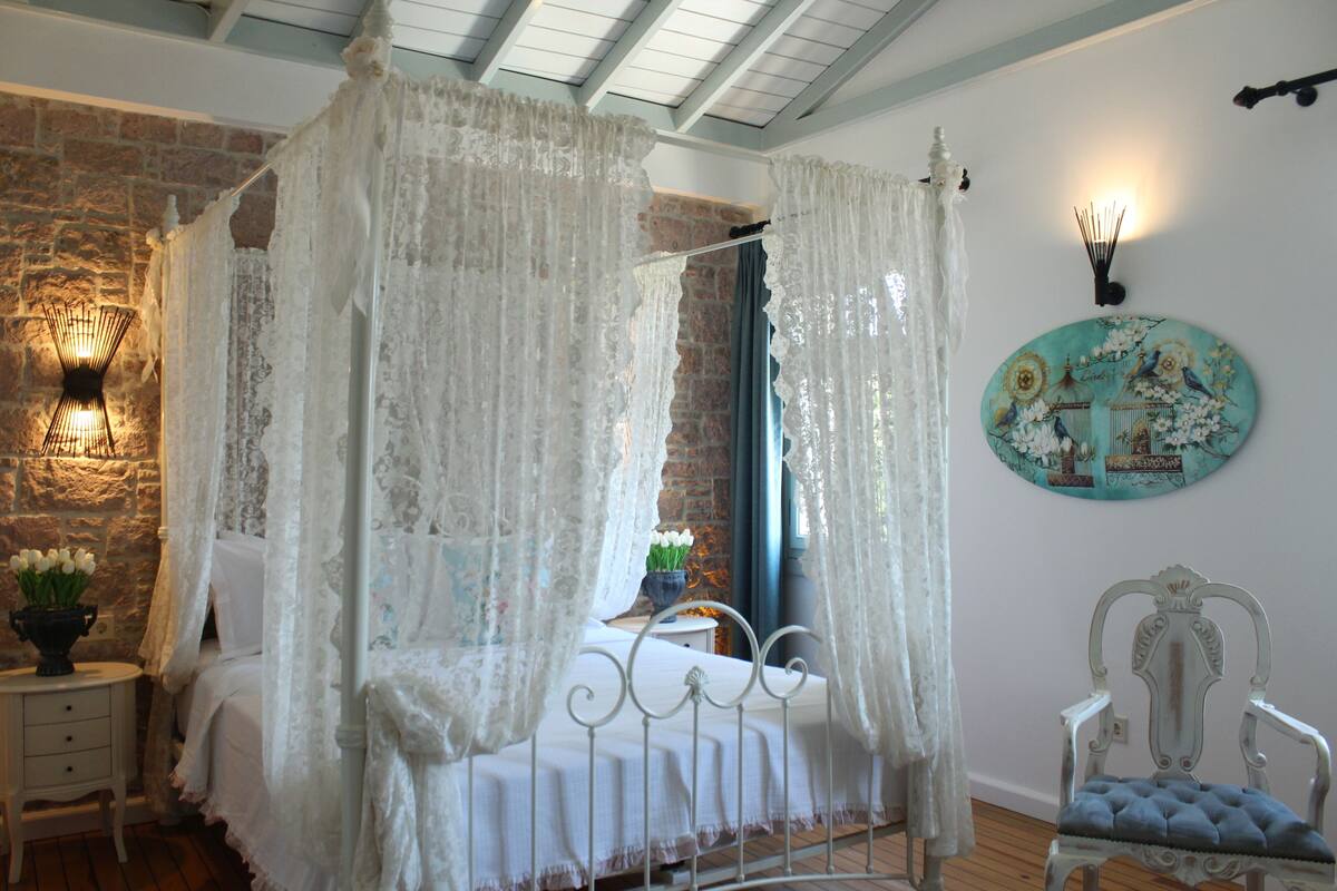 A bedroom with a four-poster bed, a stone wall, a small table with a vase of flowers, a decorative wall lamp, and a vintage chair.