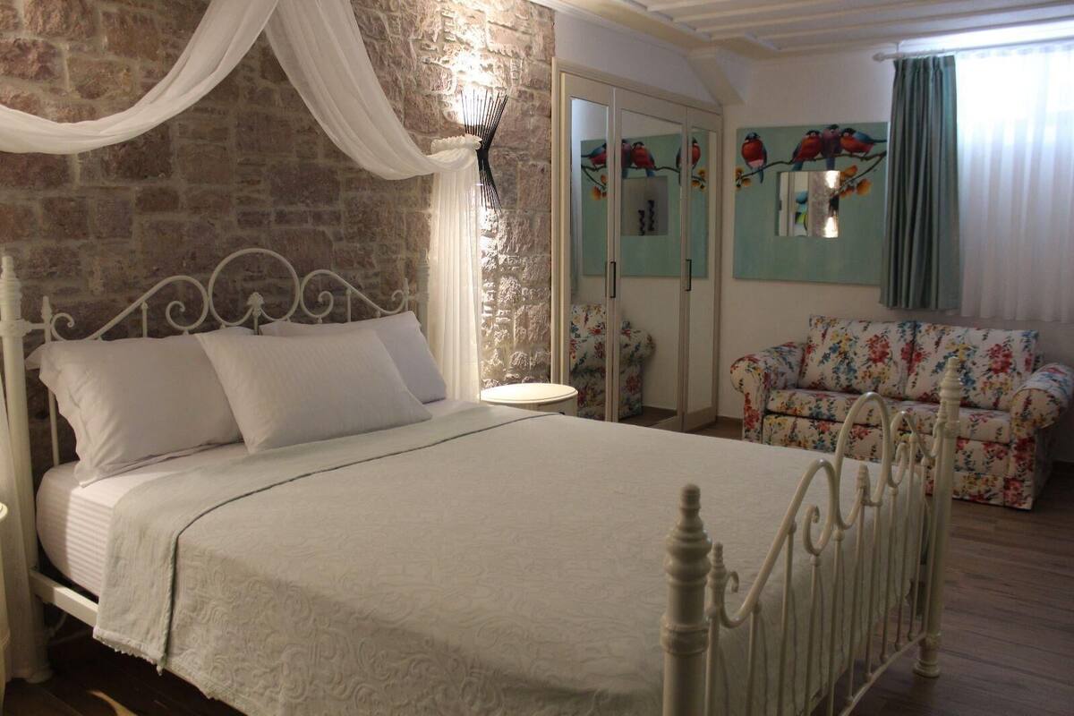 A bedroom with a white bed, a floral armchair, and a stone wall.