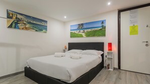 Ein ordentlich bezogenes Bett mit weißen Leinentüchern und Kissen, ein Nachttisch mit einer Lampe und zwei an der Wand hängende Bilder, die Strandansichten zeigen.