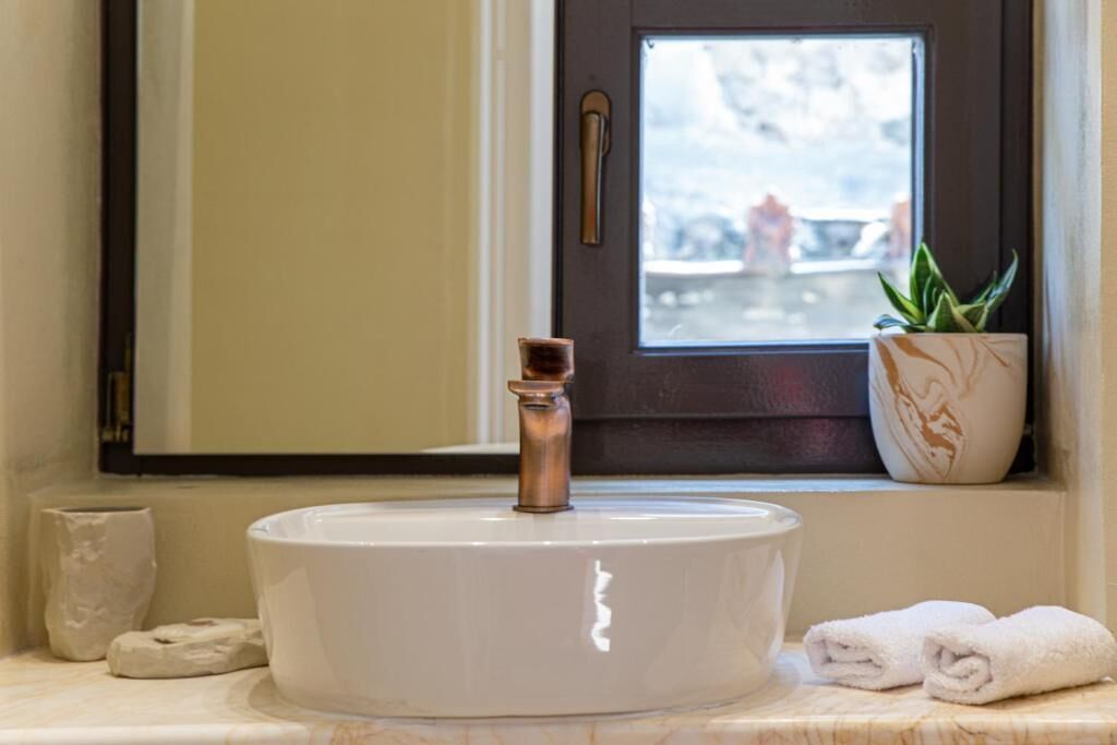 A bathroom with a round white sink, a copper faucet, a potted plant, and a window with a view of a snowy landscape.