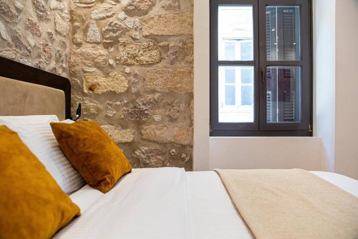A bedroom with a stone wall, a bed with white bedding and two mustard-colored pillows, and a window with blinds.