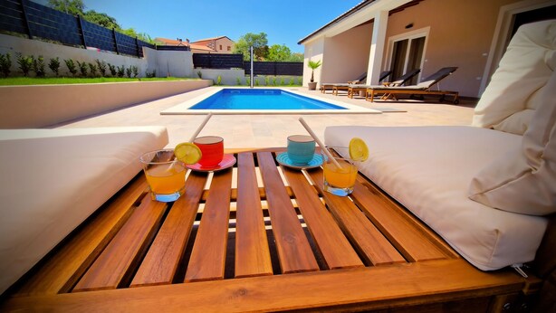 A wooden table with drinks and a glass of water, a pool, and lounge chairs in a backyard. - A picturesque holiday home, located in a quiet part of the Rovinj municipality (Rovinjsko Selo)