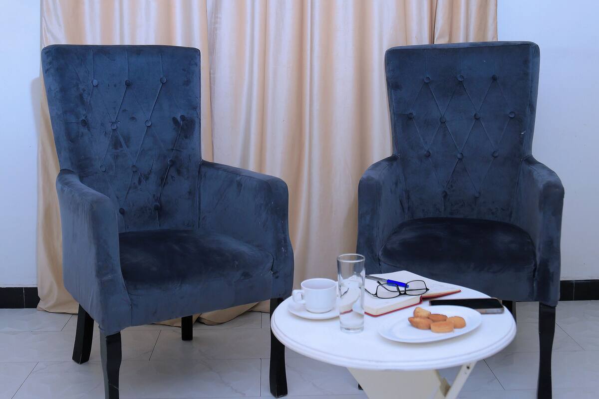 Dos sillones tapizados en azul oscuro con respaldo capitonado, una mesita redonda con mantel blanco y una cortina beige al fondo.