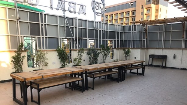 A courtyard with long wooden tables and benches, surrounded by a modern building with large windows and a metal structure. - Abat Guest House (Addis Ababa)