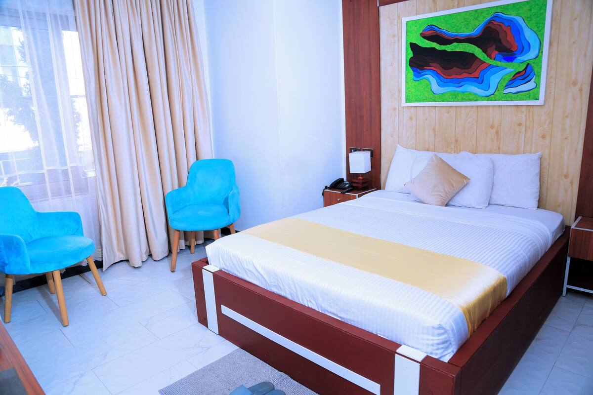 Habitación de hotel con una cama, dos sillas azules, una mesita de noche y un cuadro en la pared.