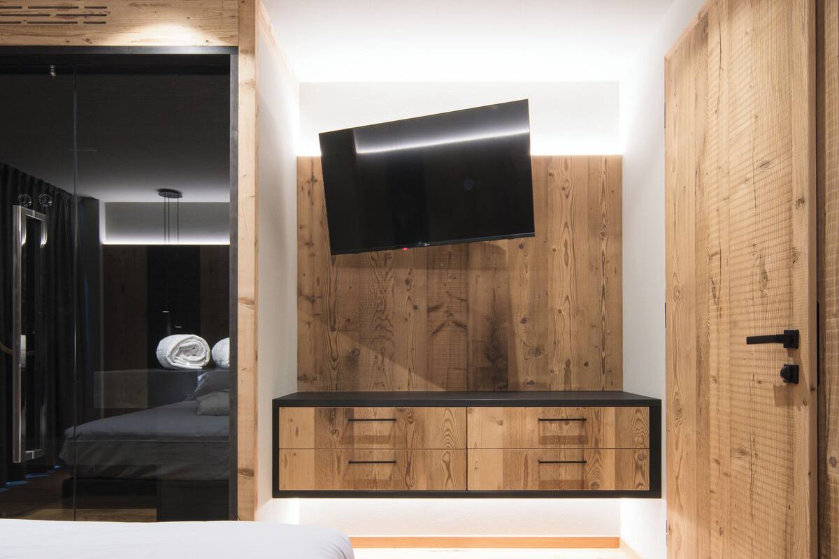 A modern bedroom with a wooden wardrobe, a flat-screen TV mounted on the wall, and a bed with white bedding.