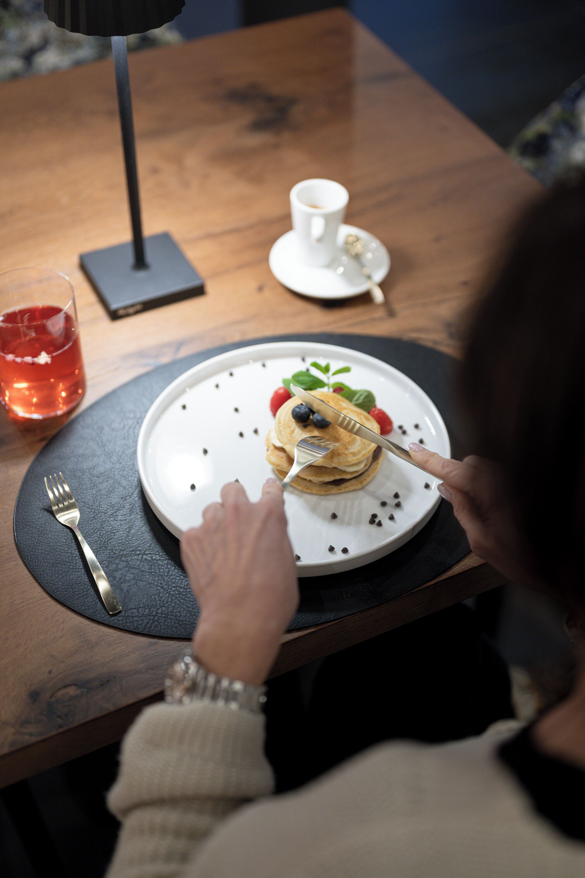 Une personne est assise à une table devant une assiette de crêpes garnies de myrtilles et de fraises, une tasse de café et un verre d’une boisson rouge.