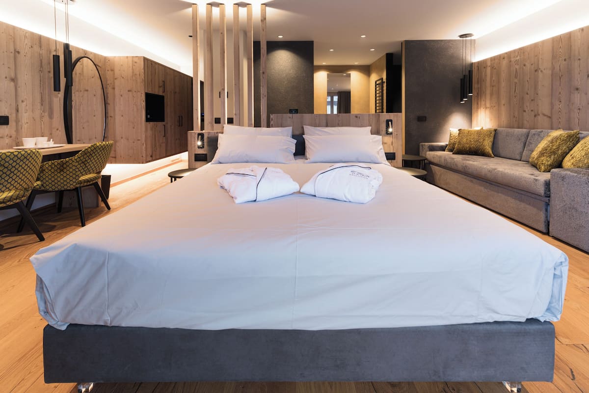A modern hotel room with a large bed, a sofa, a dining area, and wooden paneling.