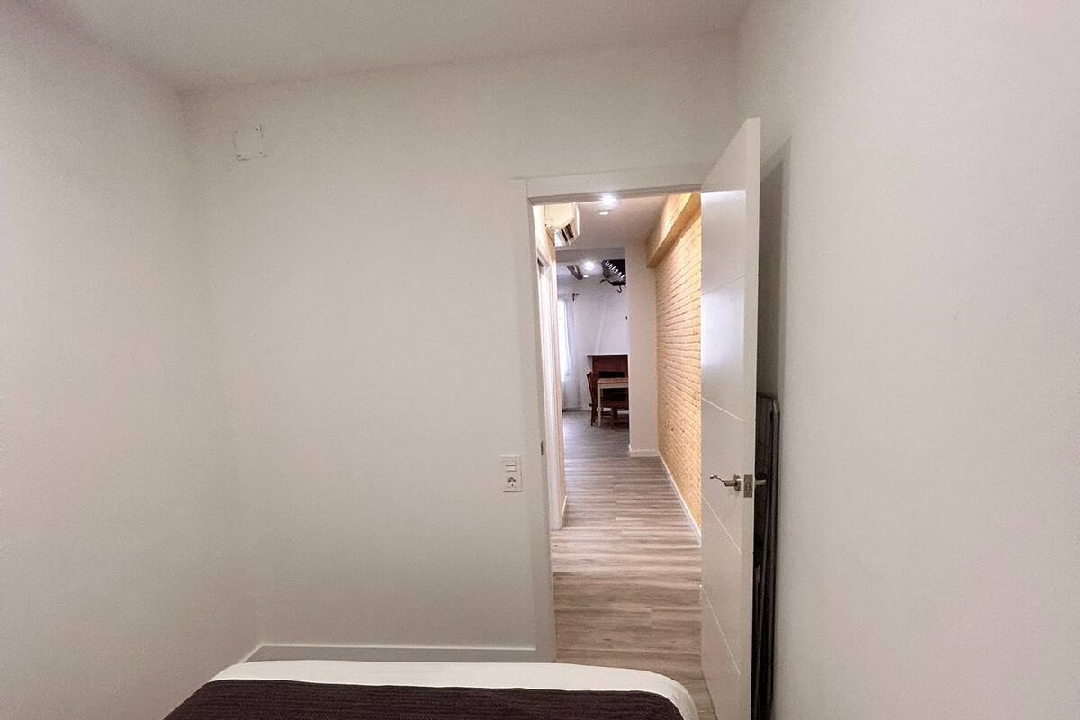 A narrow, well-lit hallway leading to a bedroom with a bed and a wooden floor.