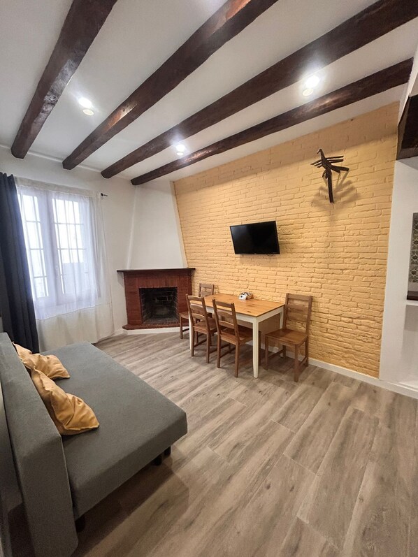 A cozy room with a fireplace, a dining table with chairs, a sofa, and a TV mounted on a brick wall. - APARTAMENTOS APARTACO (Sitges)