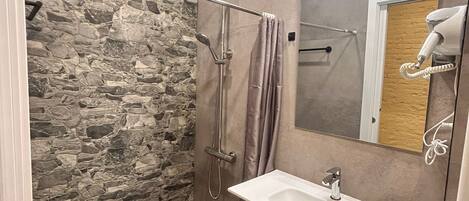 A bathroom with a stone wall, a shower with a glass door, a sink with a mirror, and a hairdryer on the wall.