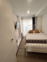 Un couloir étroit mène à un lit avec un cadre en métal, un téléviseur fixé au mur et un radiateur.