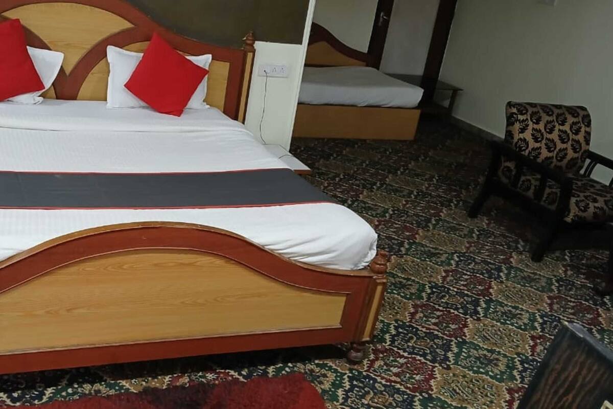 A bed with a wooden headboard, white bedding, and red pillows. There is a patterned carpet on the floor and a chair with a floral design.