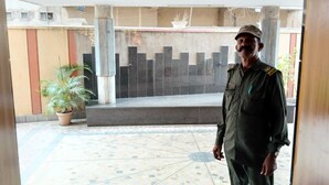 A man in uniform standing in a courtyard with a tiled floor, a potted plant, and a black gate in the background. - Hotel Sharda By Edge By GRB (Raipur)