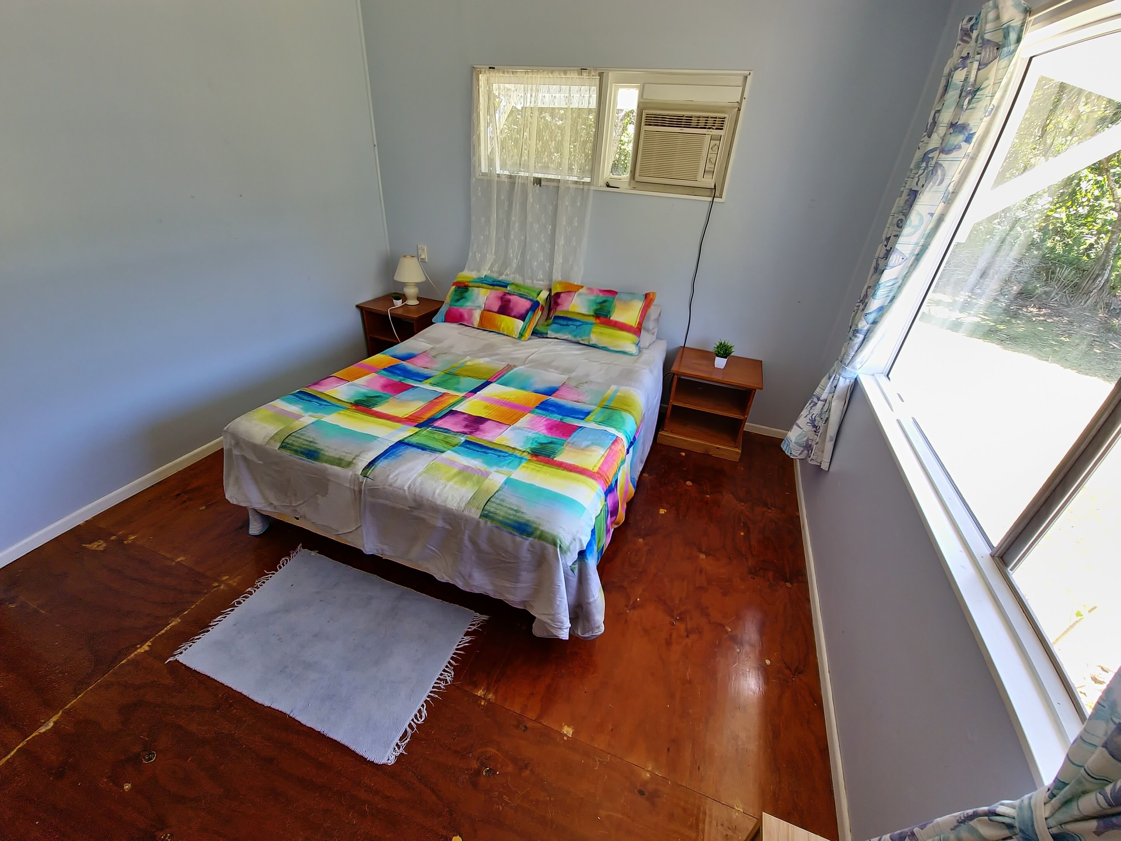 A bedroom with a bed, a nightstand, a small table with a plant, and a window with curtains.