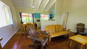 A spacious living area with a wooden floor, a wooden bench, a wooden chair, and a dining table with chairs. There is a kitchen area with a microwave and a sink. - Spatzy's Fale - Tropical Rainforest Retreat (Alofi)
