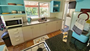 A kitchen with white cabinets, a microwave, a kettle, and a sink. There are blue shelves above the counter and a window with a view of greenery. - Spatzy's Fale - Tropical Rainforest Retreat (Alofi)