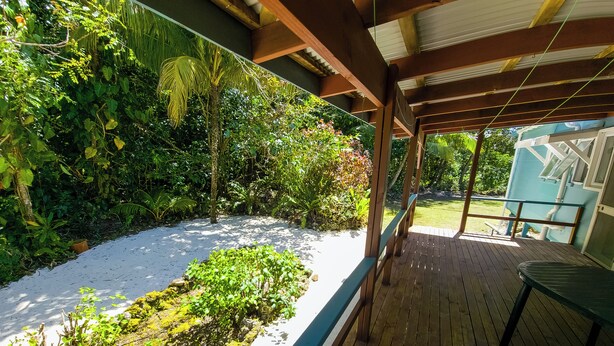 A covered outdoor area with a wooden deck, a white planter box with plants, and a view of a blue building in the background. - Spatzy's Fale - Tropical Rainforest Retreat (Alofi)