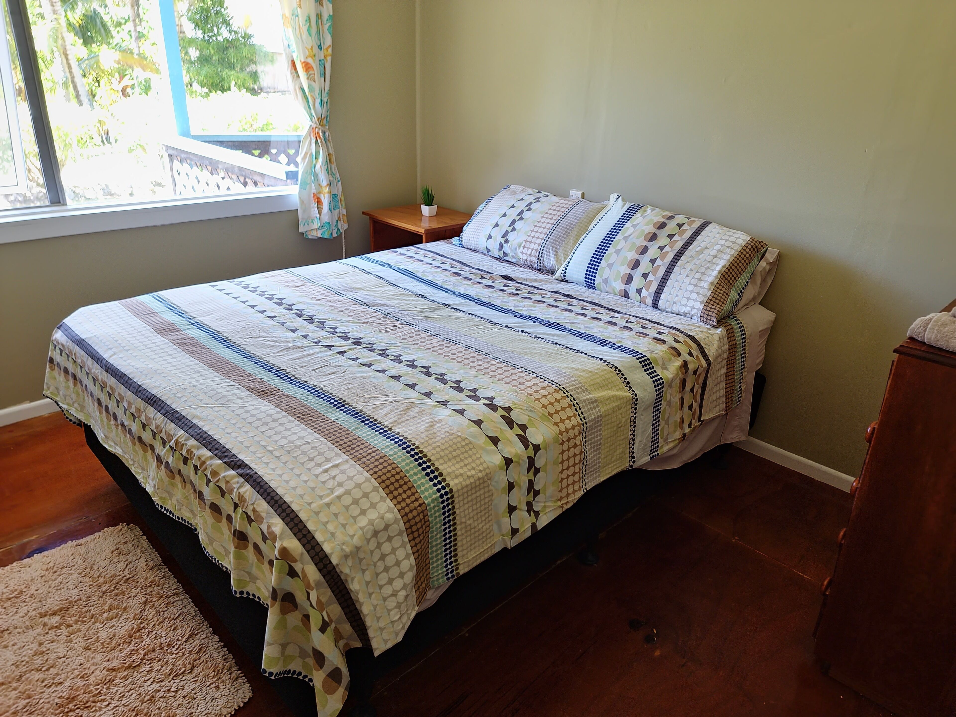 Un letto rifatto con una coperta e cuscini decorati, un comodino con una pianta e una finestra con tende.