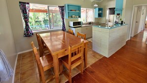 A spacious kitchen with a wooden dining table and chairs, a built-in island, and a window with curtains. - Spatzy's Fale - Tropical Rainforest Retreat (Alofi)