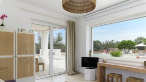 Un salon moderne doté d’un téléviseur, de meubles en bois, d’une grande fenêtre donnant sur une piscine et un jardin, ainsi que d’une porte-fenêtre coulissante qui mène à l’extérieur.