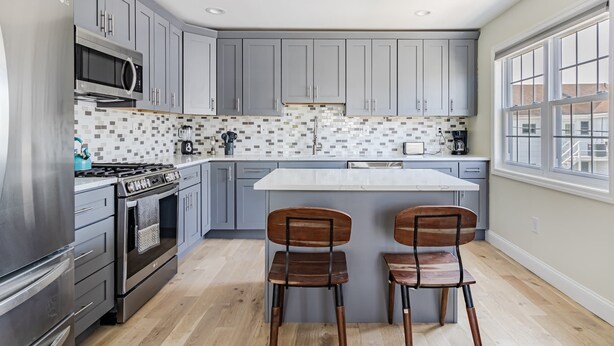 A modern kitchen with grey cabinets, a stainless steel refrigerator, a microwave, a stove, and two wooden chairs. - Spacious 3BR Condo Near Ventnor Beach (Ventnor City)