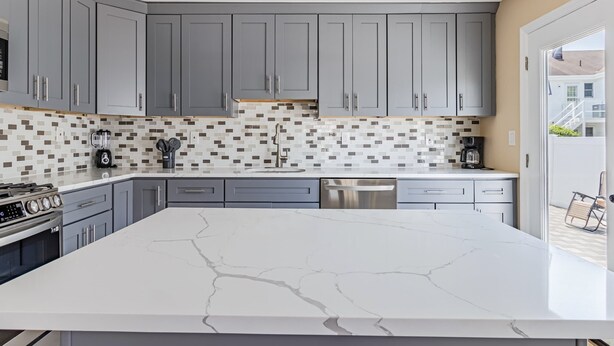 Una cocina moderna con armarios grises, una isla de mármol blanco y un salpicadero de mosaicos.