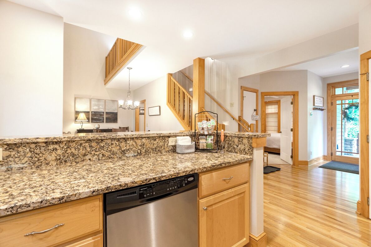 A kitchen with granite countertops, a dishwasher, wooden cabinets, a chandelier, and a staircase leading to another room.