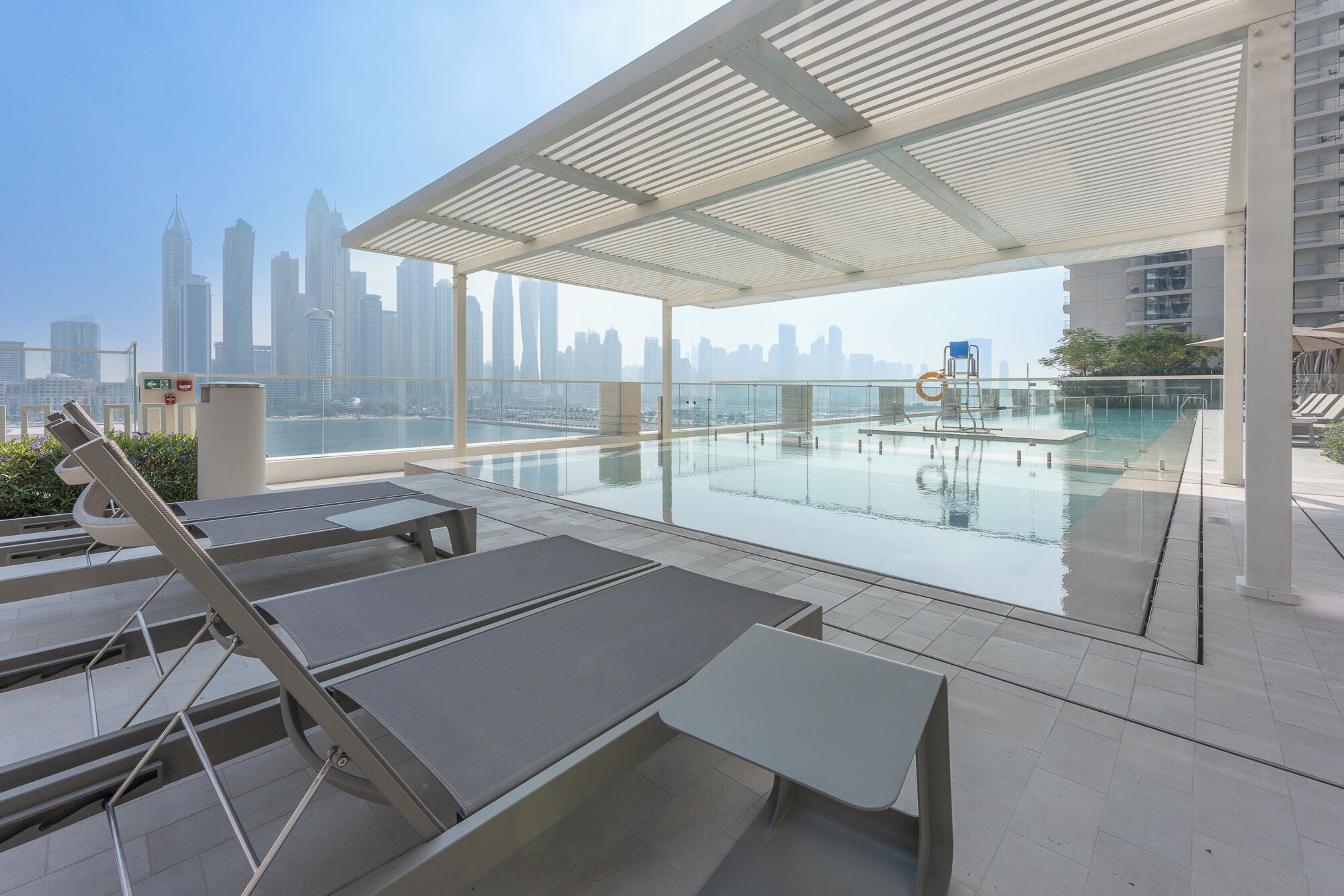 A rooftop pool area with lounge chairs and a glass barrier, overlooking a city skyline.