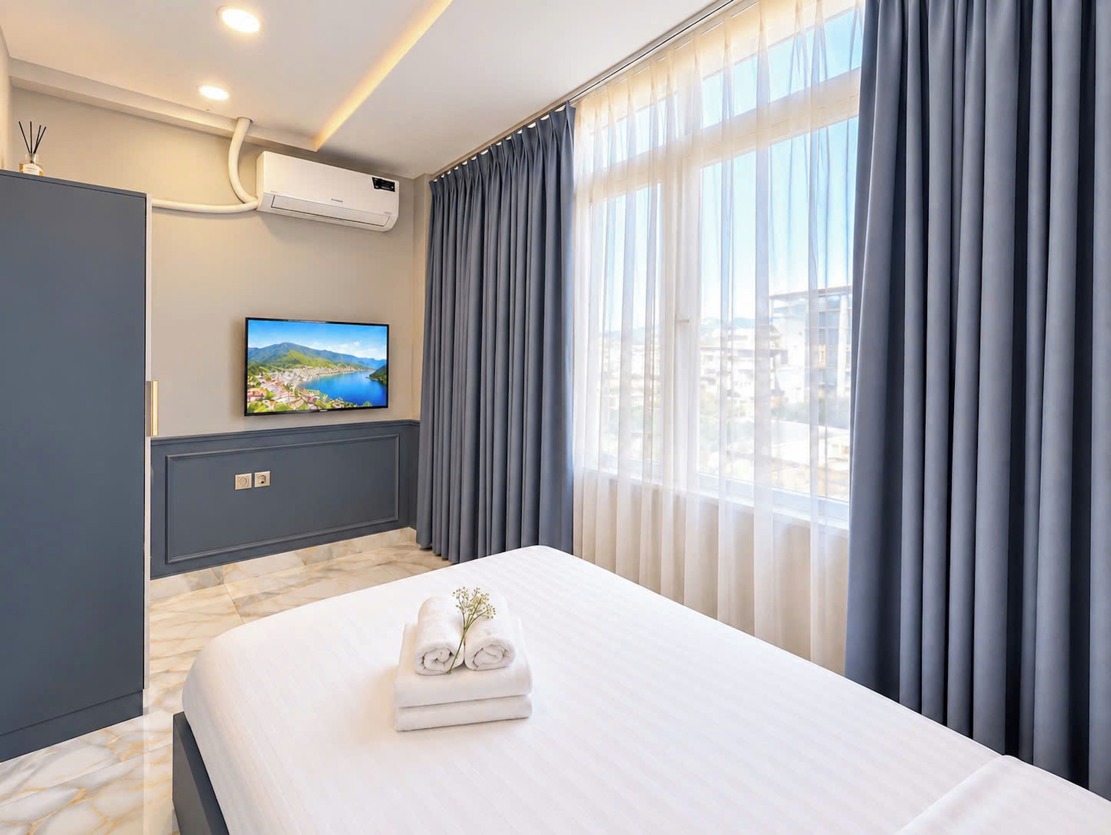A hotel room with a bed, a flat-screen TV, an air conditioning unit, and a window with curtains.
