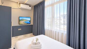 A hotel room with a bed, a flat-screen TV, an air conditioning unit, and a window with curtains.